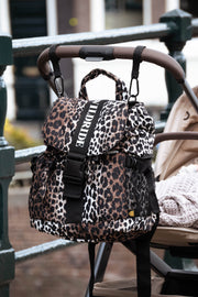 A leopard print stroller changing backpack with black straps and a prominent black strap featuring the white text 'WILDRIDE' hanging from a stroller handle in an outdoor setting.