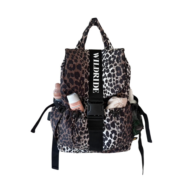 A leopard print backpack with black and white savanna print, featuring a central black strap with the brand name 'WILDRIDE' in white text, a black plastic buckle, and side pockets holding various items.