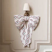 A large fabric bow with a white base, burgundy wavy lines, and small green floral motifs, hanging on a wall sconce against a beige paneled wall.