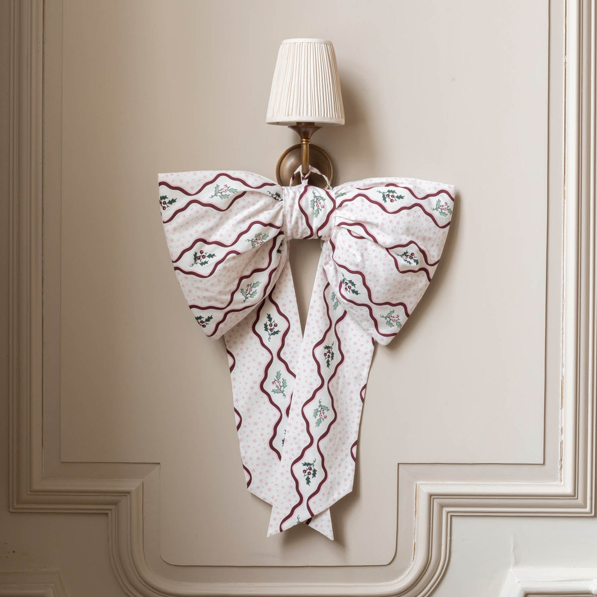 A large fabric bow with a white base, burgundy wavy lines, and small green floral motifs, hanging on a wall sconce against a beige paneled wall.