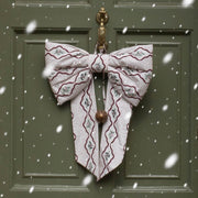 A large decorative bow made from patterned fabric hangs on a dark green front door. The bow has a white base with burgundy wavy lines and small gray floral motifs, plus a small metallic bell at the center.