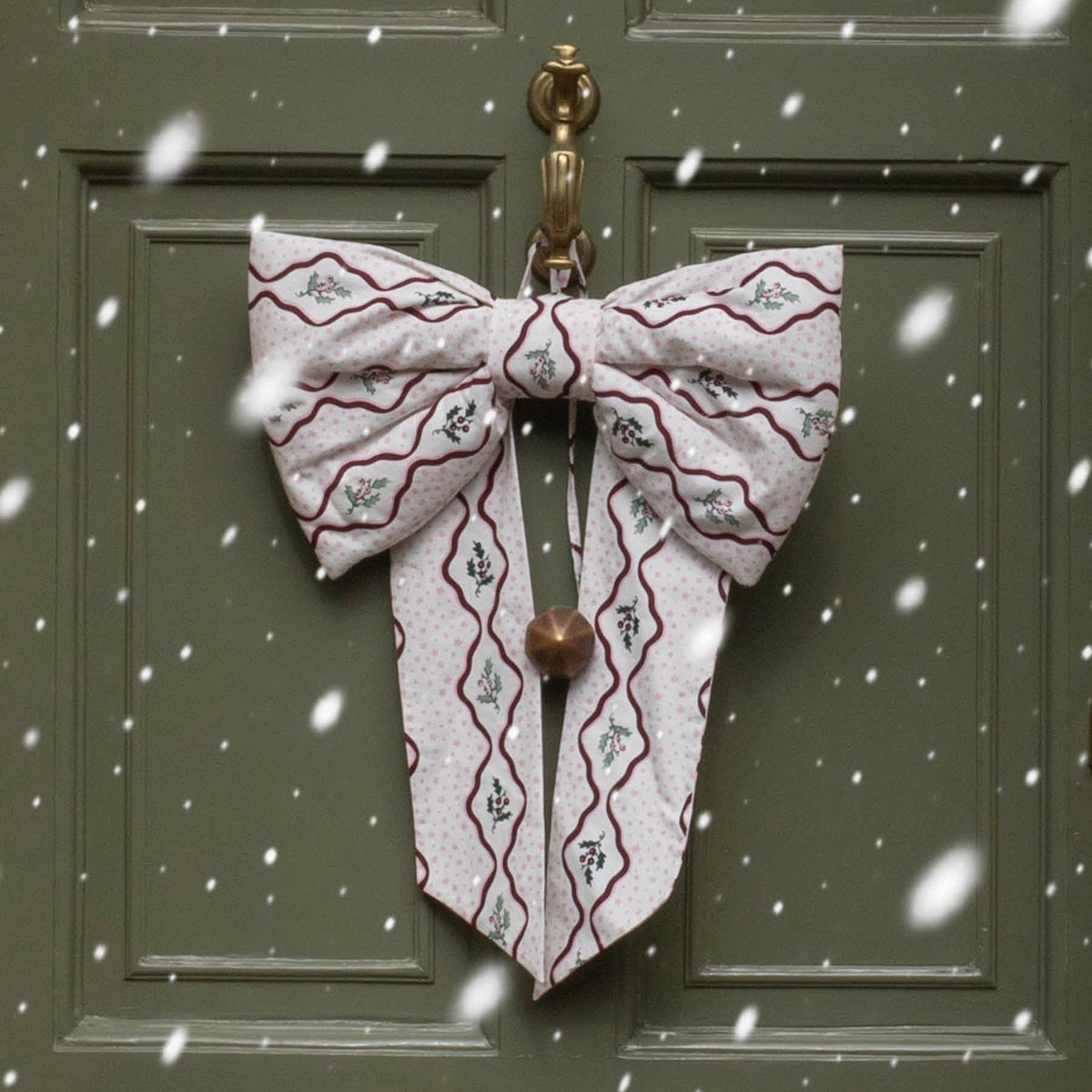A large decorative bow made from patterned fabric hangs on a dark green front door. The bow has a white base with burgundy wavy lines and small gray floral motifs, plus a small metallic bell at the center.