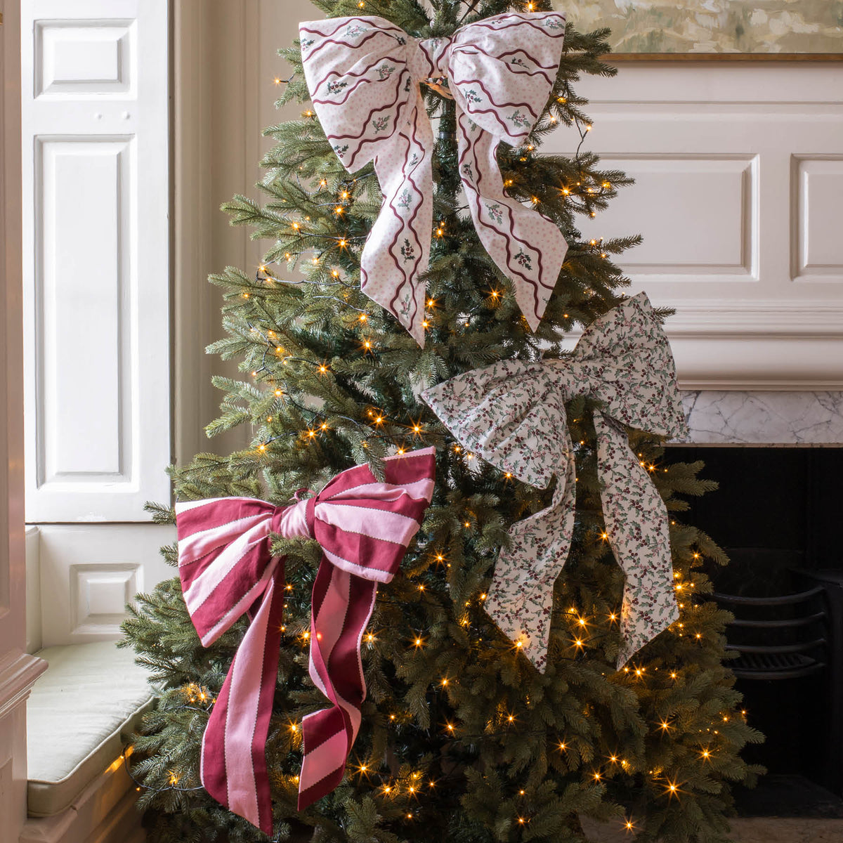 A decorated Christmas tree in a fireplace room, featuring two large decorative bows: one in pink and white stripes and one in a black and white floral pattern, with warm white string lights throughout.