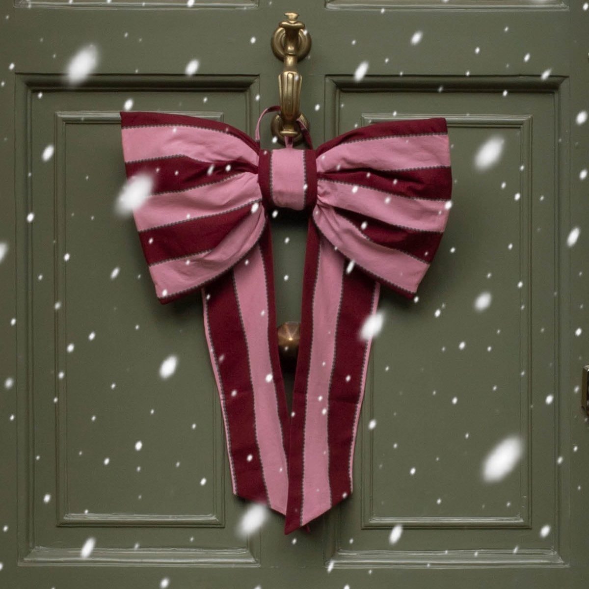 A large decorative bow made from pink and burgundy striped fabric hangs on the brass doorknob of an olive green front door, with falling snow visible in the scene.