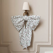 A large fabric bow with a delicate white, green, and red floral print hangs on a beige paneled wall below a wall sconce.