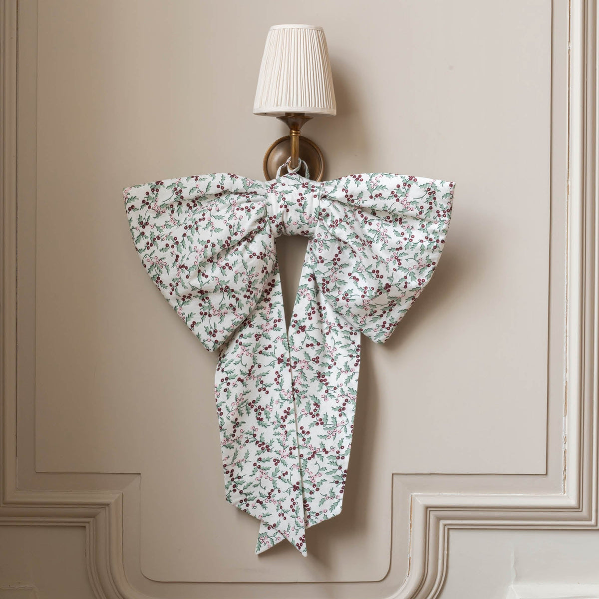 A large fabric bow with a delicate white, green, and red floral print hangs on a beige paneled wall below a wall sconce.