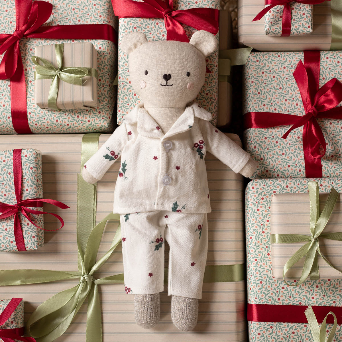 A plush teddy bear wearing Christmas pyjamas lies centered among wrapped gift boxes with red and green ribbons on a striped background.
