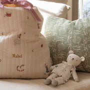 A cream quilted Christmas sack with embroidered sugar plum fairy motifs, personalised with the name 'Mabel' in gold, styled on a sofa next to a green patterned pillow and a teddy bear wearing Christmas pyjamas