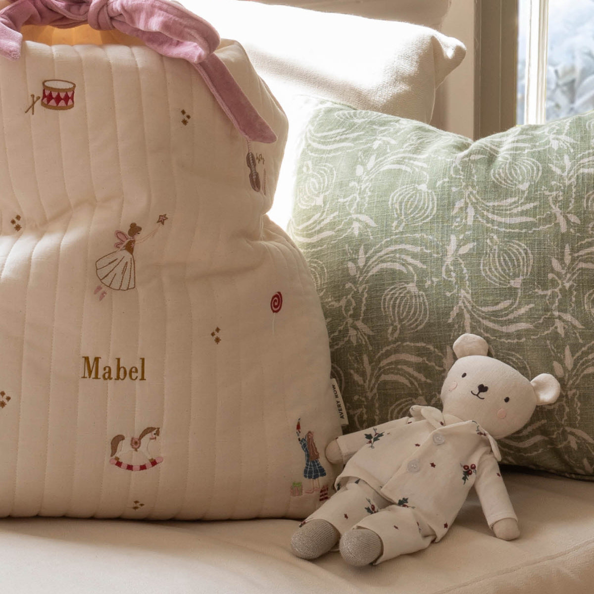 A cream quilted Christmas sack with embroidered sugar plum fairy motifs, personalised with the name 'Mabel' in gold, styled on a sofa next to a green patterned pillow and a teddy bear wearing Christmas pyjamas
