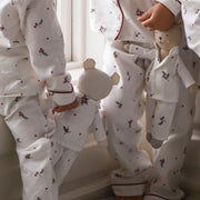 Three children wearing matching white long-sleeve pajamas with a small red and green floral print and red piping trim, standing barefoot indoors near a window.