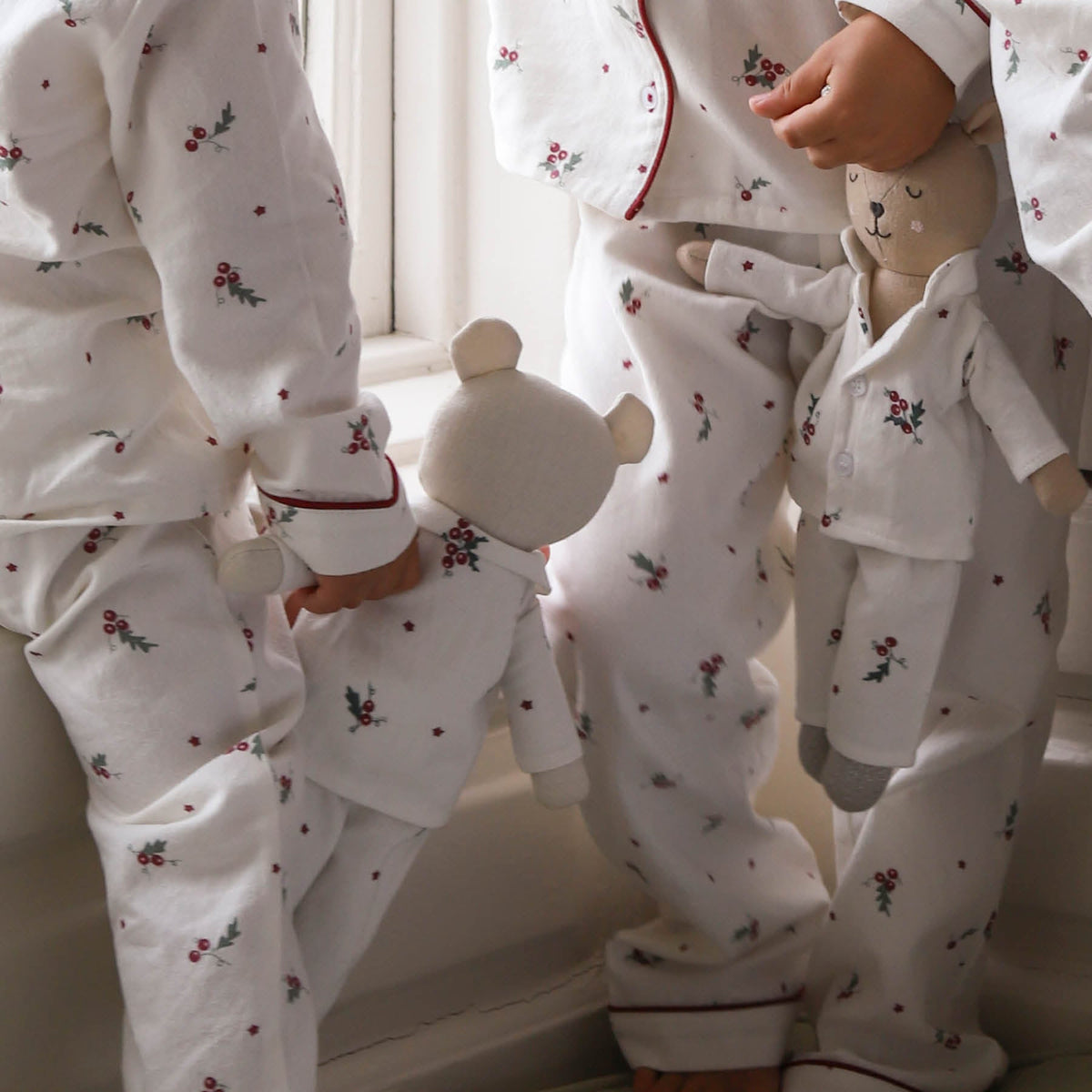 Three children wearing matching white long-sleeve pajamas with a small red and green floral print and red piping trim, standing barefoot indoors near a window.