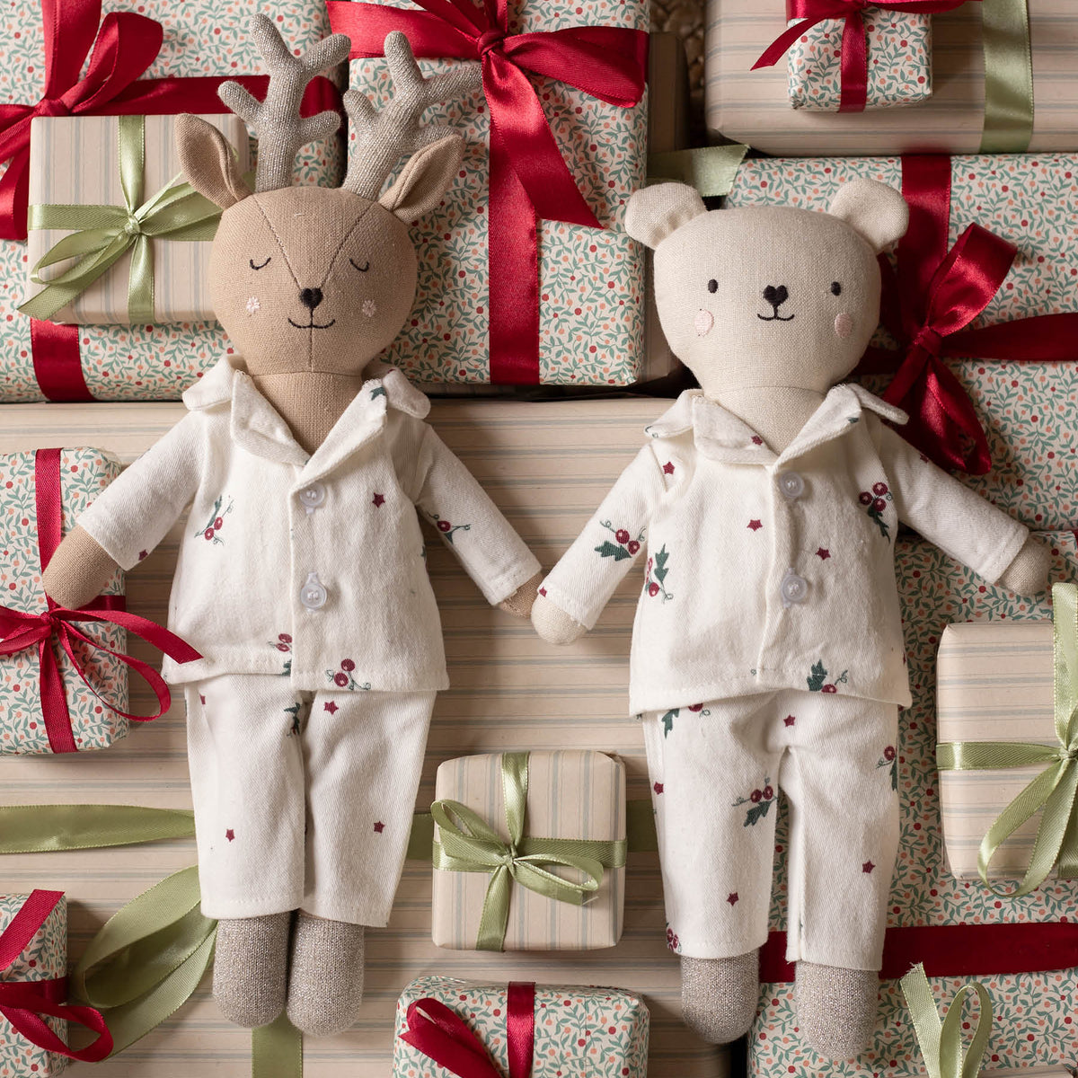 Two plush stuffed toys dressed in matching Christmas pyjamas, surrounded by wrapped gift boxes with red and green ribbons. The left toy is a reindeer with antlers, and the right toy is a bear, both in cream pajamas with small floral and star prints.