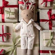 A plush deer doll wearing Christmas pyjamas, posed among wrapped gift boxes with red and green ribbons and patterned paper.