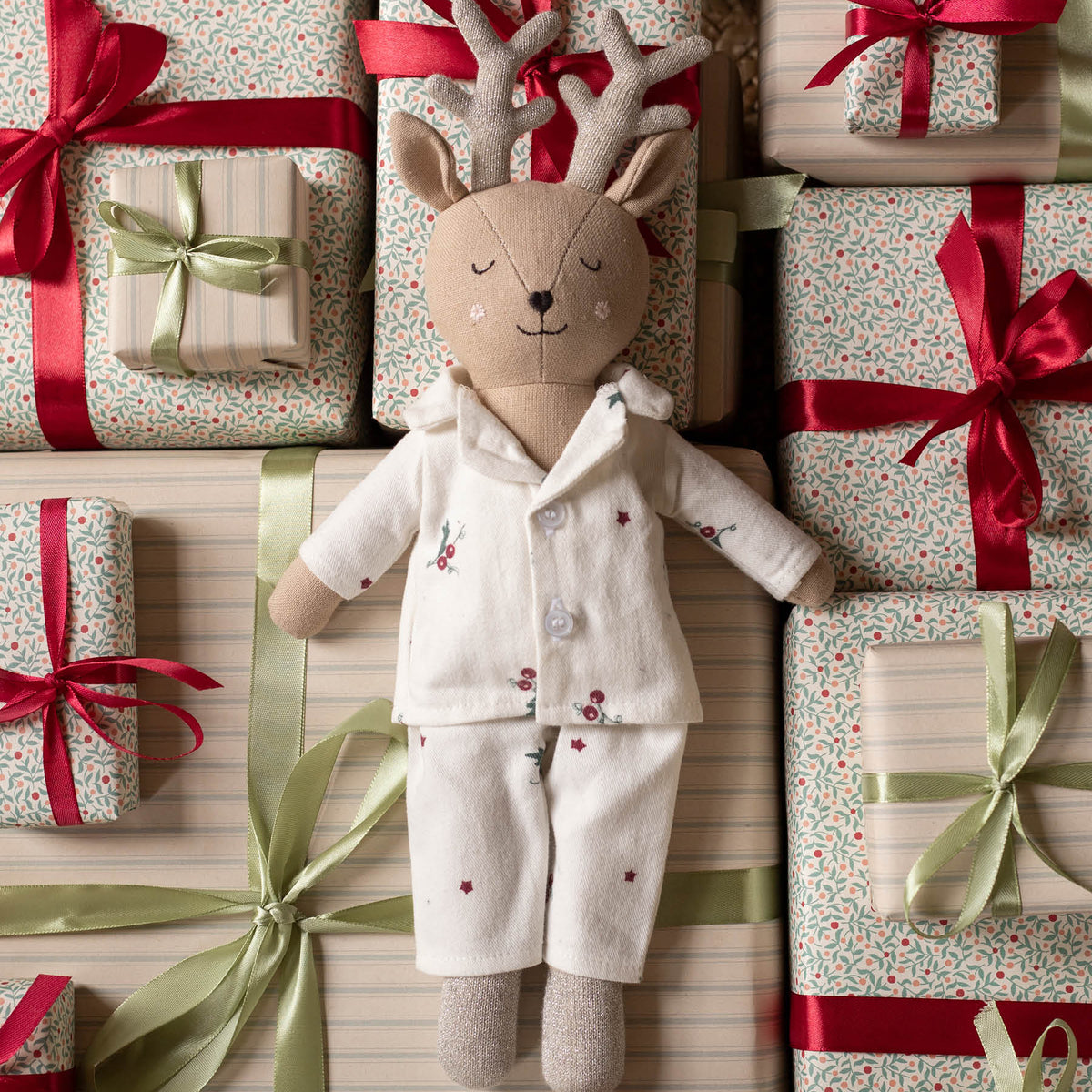 A plush deer doll wearing Christmas pyjamas, posed among wrapped gift boxes with red and green ribbons and patterned paper.