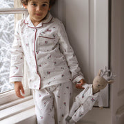 A young child wearing a Christmas pyjama set with red piping and holly print, standing by a window and holding a stuffed reindeer doll dressed in matching pyjamas.