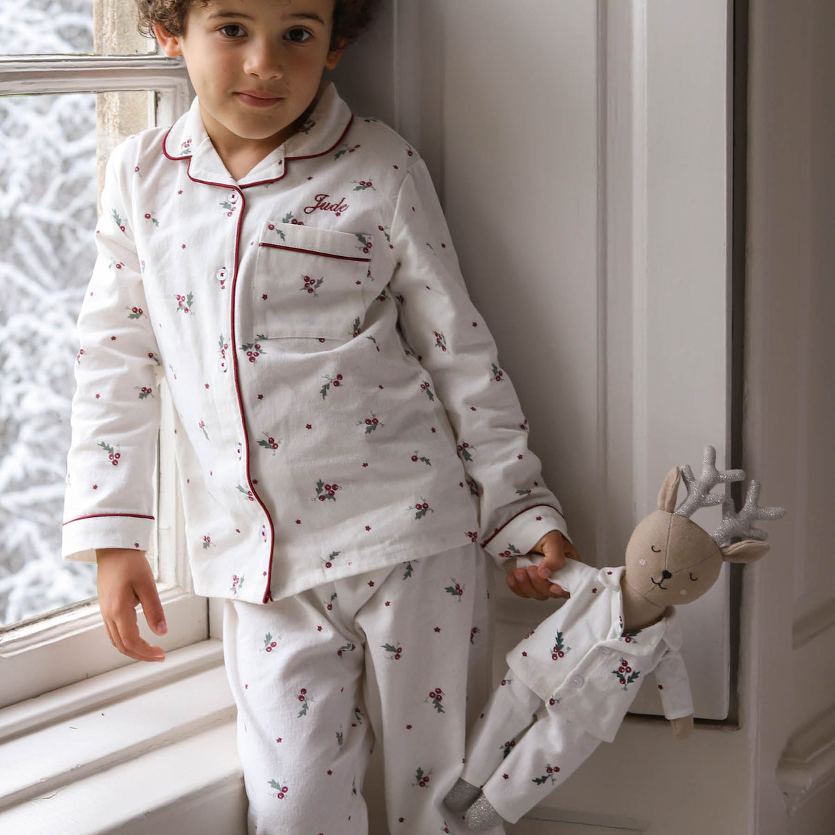 A young child wearing a Christmas pyjama set with red piping and holly print, standing by a window and holding a stuffed reindeer doll dressed in matching pyjamas.