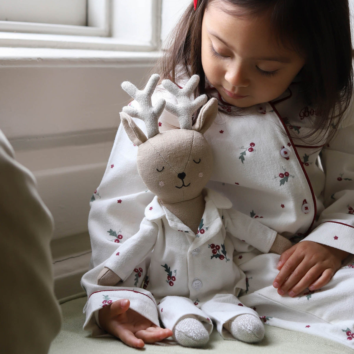 A young child wearing white Christmas-themed pyjamas sits indoors next to a matching plush reindeer stuffed toy wearing matching pyjamas with soft natural light coming from a nearby window.