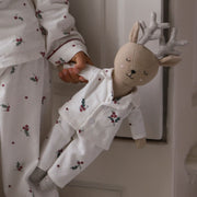 A child wearing white holiday-themed pyjamas with a red berry and holly print, holding a plush stuffed deer doll dressed in matching white pyjamas with glittery silver antlers.