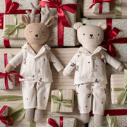 Two plush stuffed toys dressed in matching holiday pyjamas, surrounded by wrapped gift boxes with red and green ribbons. The left toy is a reindeer with antlers, and the right toy is a bear, both in cream pajamas with small floral and star prints.