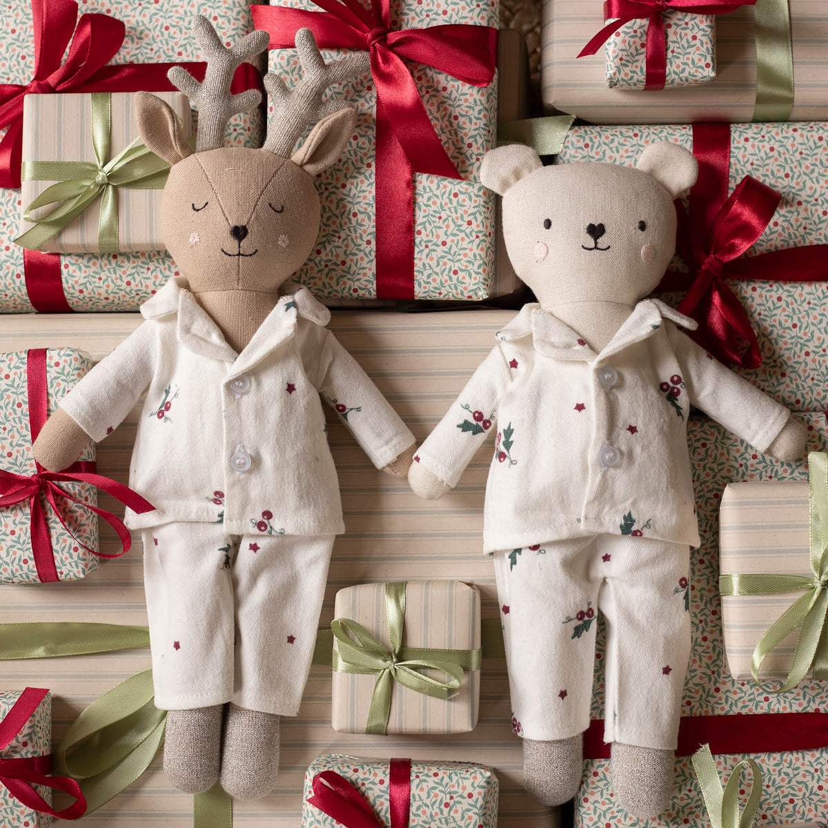Two plush stuffed toys dressed in matching holiday pyjamas, surrounded by wrapped gift boxes with red and green ribbons. The left toy is a reindeer with antlers, and the right toy is a bear, both in cream pajamas with small floral and star prints.