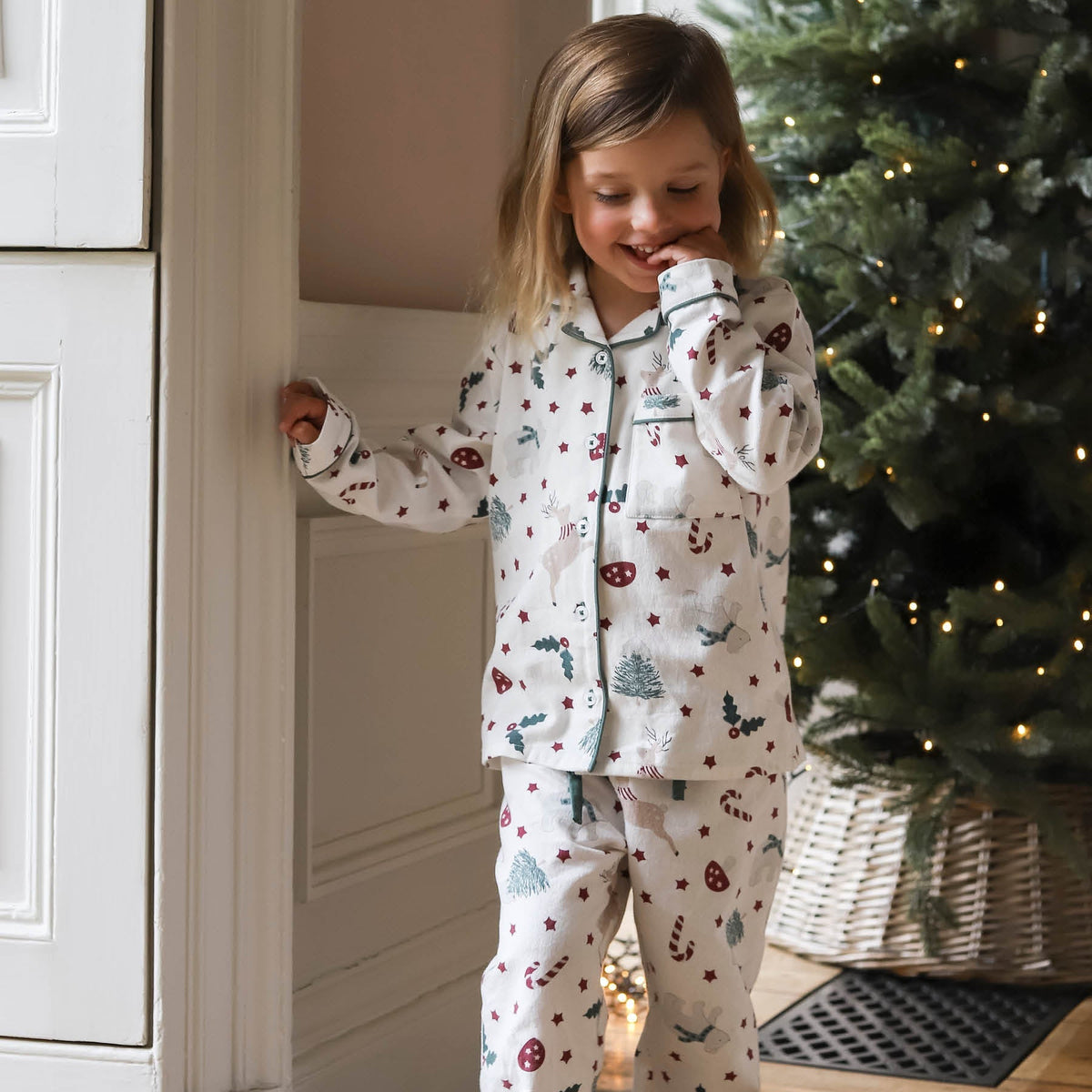 A young girl wearing a long-sleeve pyjama set with a Christmas print, standing in a cosy room next to a white paneled wall and a decorated Christmas tree.