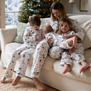 A family of four wearing matching Christmas pyjamas sits together on a cream-colored sofa in a cosy living room with a decorated Christmas tree in the background.