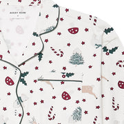 A close-up of a pyjama top with a festive Christmas print on an off-white background. The pattern features dark green holly leaves, red candy canes, red stars, beige reindeer, and green Christmas trees.