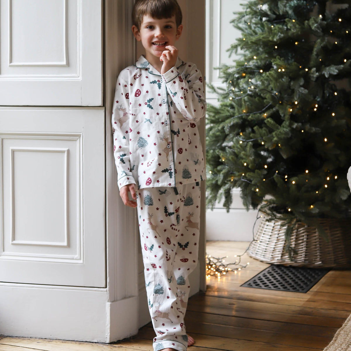 A young boy wearing a white long-sleeve pyjama set with a colorful Christmas tree and snowman print, standing barefoot in a doorway next to a decorated Christmas tree.
