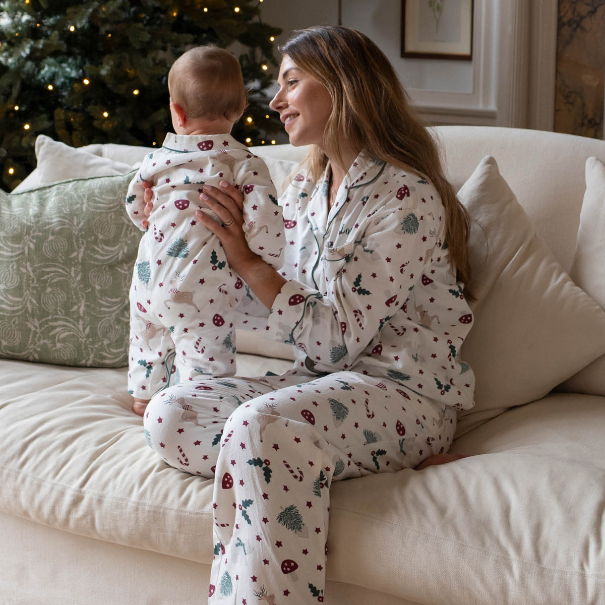 A woman and a baby wearing matching Christmas pyjamas sit together on a cream-colored couch in a cosy living room with a Christmas tree in the background.