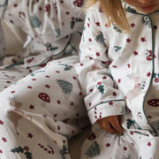 A close-up of a child wearing a cosy white Christmas-themed pyjama set with red stars, green pine trees, candy canes, and reindeer motifs. The soft fabric and festive print are shown in a warm, cosy indoor setting.