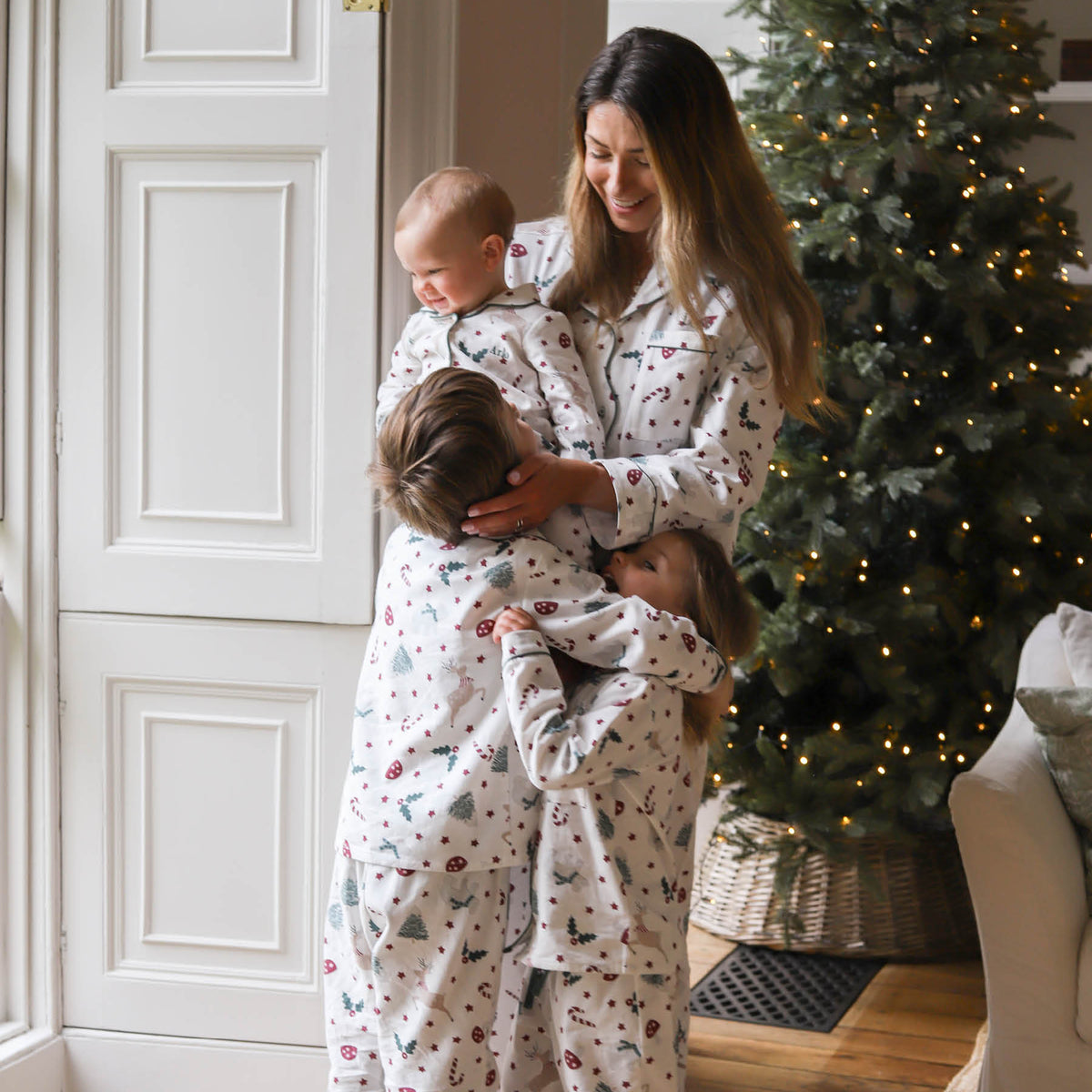 Three children and a woman wearing matching white Christmas-themed pyjamas hugging in a cosy living room with a lit Christmas tree in the background.