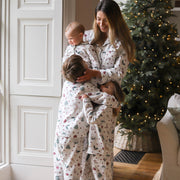 A woman holding a baby and standing with two children wearing matching Christmas-themed pyjamas with a festive print, standing in a cosy room next to a decorated Christmas tree.