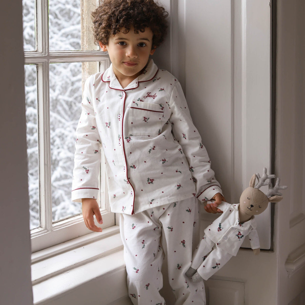 A young child wearing a white long-sleeve pyjama set with a holly leaves and berries pattern, leaning against a window frame and holding a reindeer soft toy.