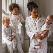 A family of four wearing matching white long-sleeve pyjamas with a small holly leaves and berries pattern and red piping. The set includes a matching adult pyjama set worn by a mother holding a baby, a toddler standing by a window, and a baby sitting on a window sill.