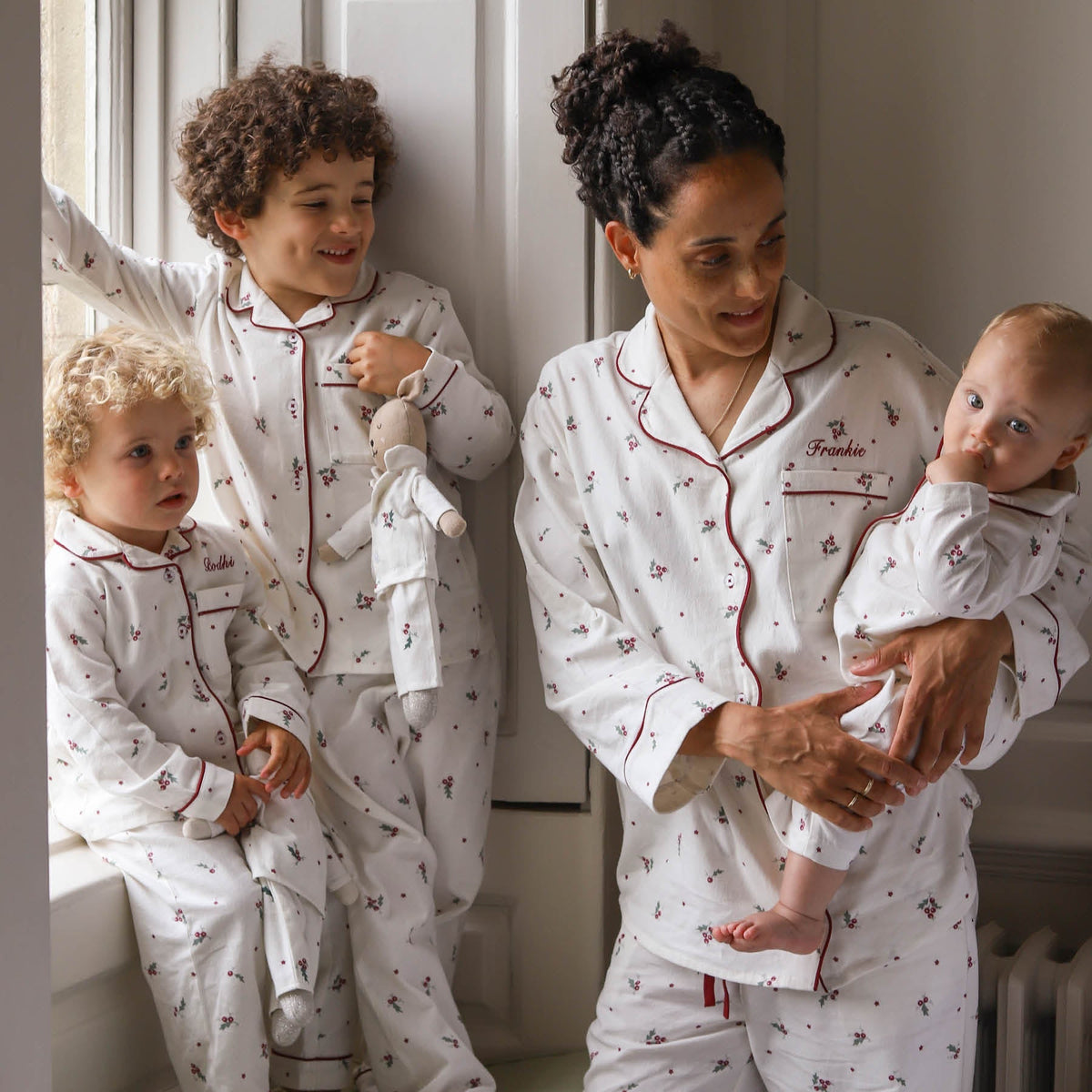 A family of four wearing matching white long-sleeve pyjamas with a small holly leaves and berries pattern and red piping. The set includes a matching adult pyjama set worn by a mother holding a baby, a toddler standing by a window, and a baby sitting on a window sill.