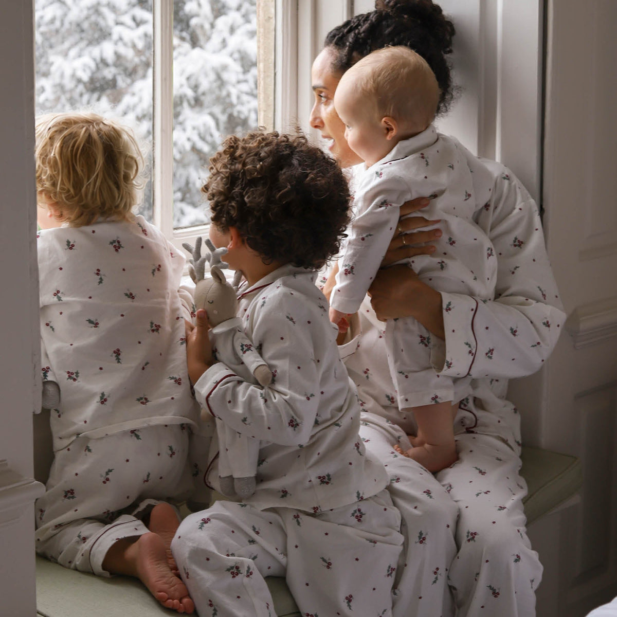 A family of four wearing matching Christmas pyjamas, sitting by a window with snow-covered trees outside. The adults and children are shown in a cosy indoor setting.