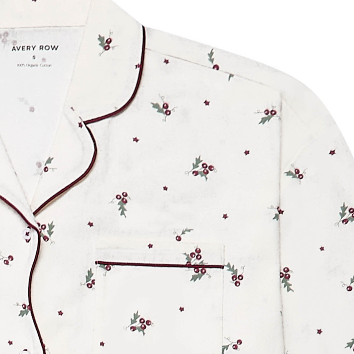 A close-up of a white long-sleeve pyjama top by Avery Row, featuring a delicate all-over print of small red berries with green leaves and dark burgundy piping along the collar, placket, and pocket.
