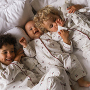 Three children lying on a bed wearing matching white long-sleeve pyjamas with a small red and green floral print and dark red piping. One child is holding a white teddy bear.