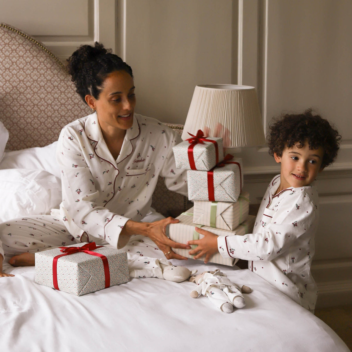 A woman and a young boy wearing matching white Christmas-themed pyjamas with a holly leaves and berries pattern, sitting on a bed with wrapped gift boxes and a stack of presents.