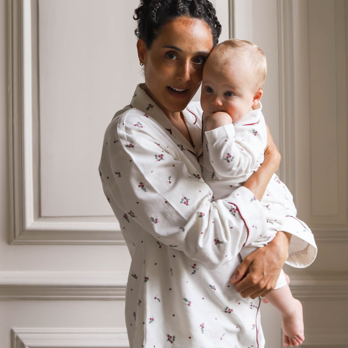 A woman holding a baby, both wearing matching white long-sleeve Christmas pyjamas with a holly leaves and berries pattern, posed against a light beige paneled wall.