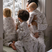 A family of four wearing matching white Christmas pyjamas with a holly pattern, sitting by a window with snow-covered trees outside. The adults and children are shown in a cosy indoor setting.