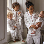 A family wearing matching Christmas pyjamas, including a woman holding a baby, a standing child, and a toddler by a window.