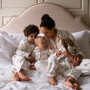 A family of four wearing matching Christmas pyjamas sits together on a bed with white linens and a patterned upholstered headboard.