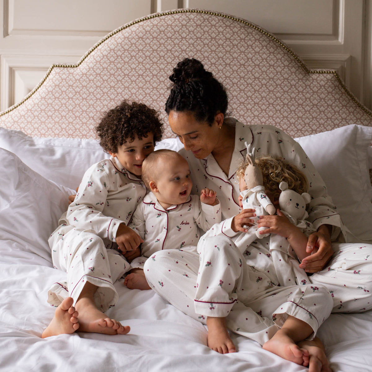 A family of four wearing matching Christmas pyjamas sits together on a bed with white linens and a patterned upholstered headboard.