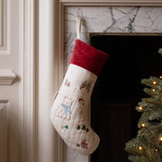 A cream-colored Christmas stocking with embroidered polar bear motifs and a red velvet cuff, hanging in front of a marble fireplace next to a lit Christmas tree.