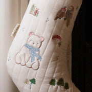 A close-up of a quilted Christmas stocking in cream with embroidered woodland motifs, including a polar bear in a blue scarf, an owl, gifts, mushrooms, and trees. A red cuff is visible at the top, and a small fabric tag is attached near the left side.