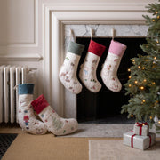 Four Christmas stockings with festive prints are displayed around a fireplace. Three stockings hang from the mantel, while two more are placed on the floor beside a lit Christmas tree and wrapped gift boxes.