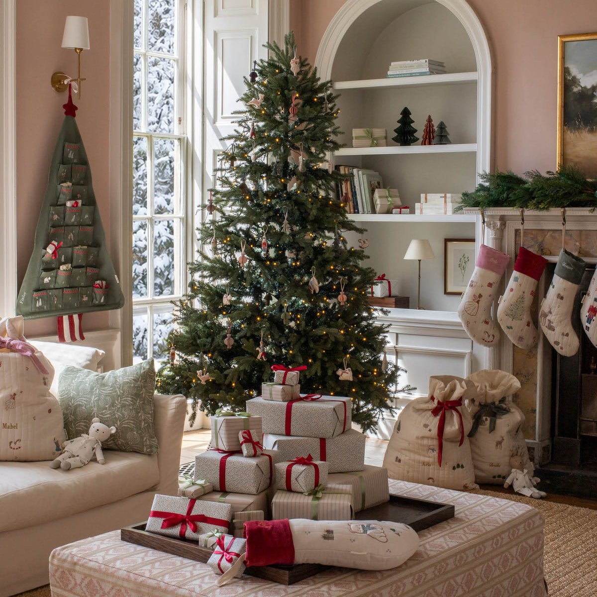 A beautifully decorated Christmas living room featuring a tall green tree with ornaments, wrapped gift boxes on a patterned ottoman, stockings hanging from a fireplace, and a cozy armchair.