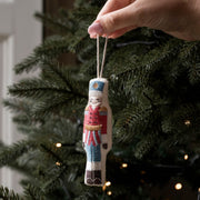 A hand holds a small embroidered nutcracker Christmas ornament against the green branches of a lit Christmas tree.