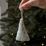 A hand holds a small triangular embroidered Christmas tree ornament with a white string hanger, featuring blue, green, and gold thread details, set against a blurred Christmas tree background with warm lights.